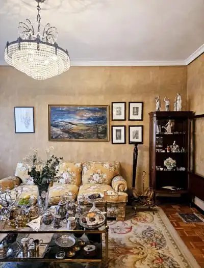 Mercadillo PL. Reyes Magos Una sala de estar vintage con paredes marrones cuenta con un sofá con estampado floral, una mesa de café de vidrio decorada con baratijas y jarrones, cuadros enmarcados en la pared, una lámpara de araña encima y un estante de madera que exhibe figuras y platos.