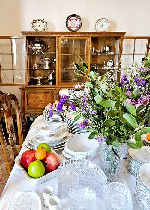 Una mesa de comedor está dispuesta con porcelana blanca, cristalería y un centro de mesa con flores de colores. Cerca, se colocan frutas variadas, incluidas manzanas y naranjas. En el fondo, un armario de madera muestra más porcelana y cubertería.